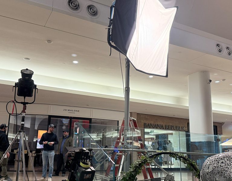 Large film lights and camera equipment are set up inside a shopping centre near various shop entrances, with two people standing nearby and a ladder in the background.