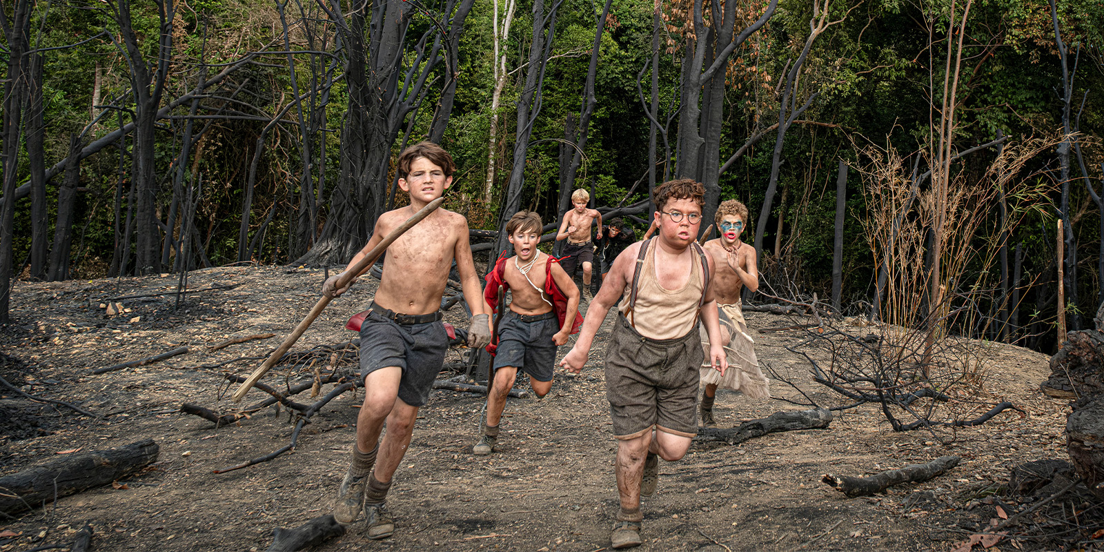 Six boys, barefoot and shirtless, run on a dirt path through a burnt forest. Their skin and clothes are dirty, and they appear determined or distressed. The background shows charred trees and green foliage.