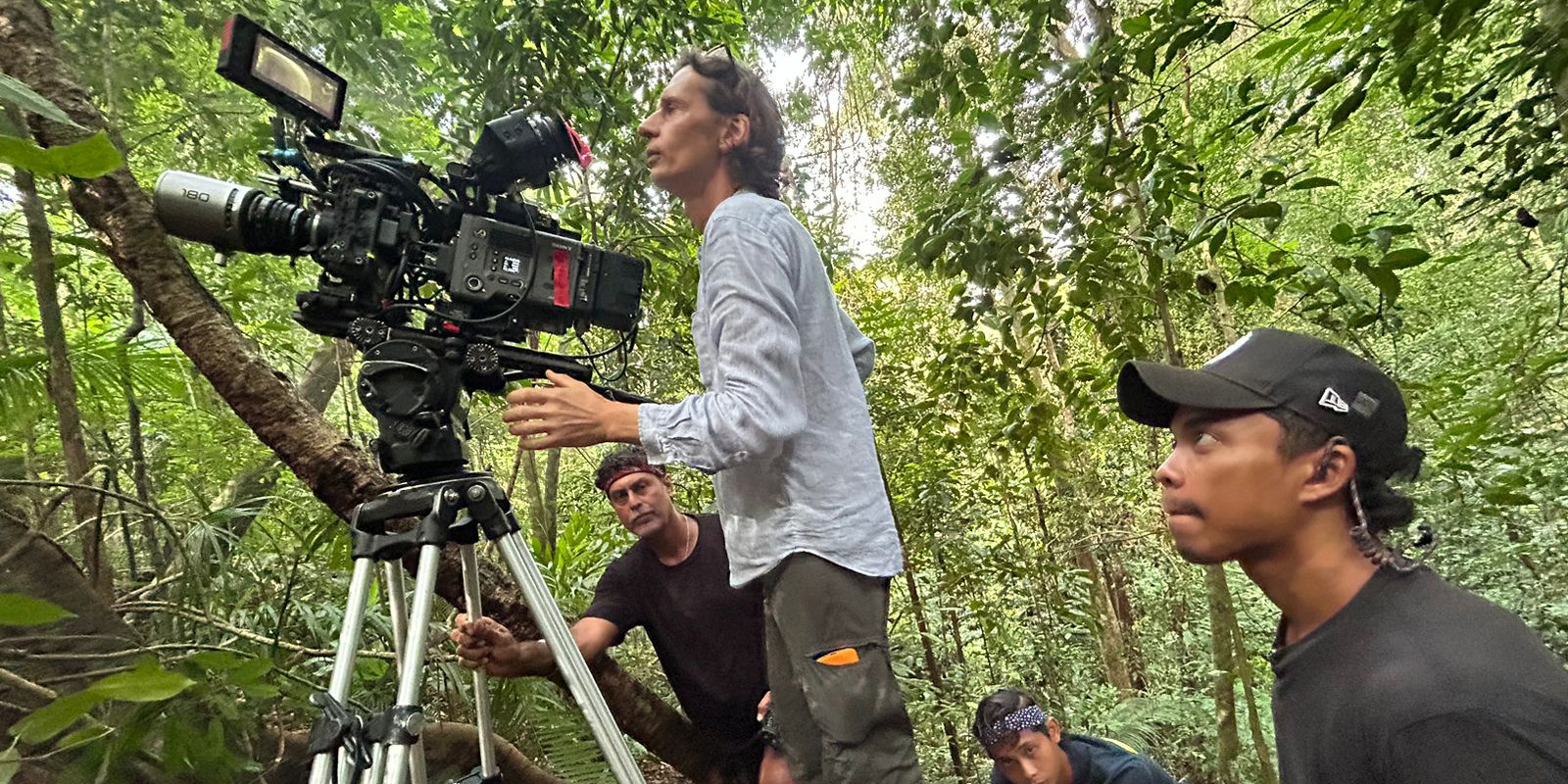 A group of people in a forest set up and operate a professional video camera on a tripod. One person adjusts the camera while others observe or assist. Dense green foliage surrounds them.