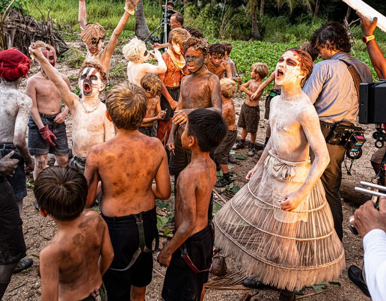 A group of children with painted faces and bodies, some in tattered clothes, gather outdoors on a dirt ground. A film crew with camera equipment captures the scene; trees and greenery are in the background.