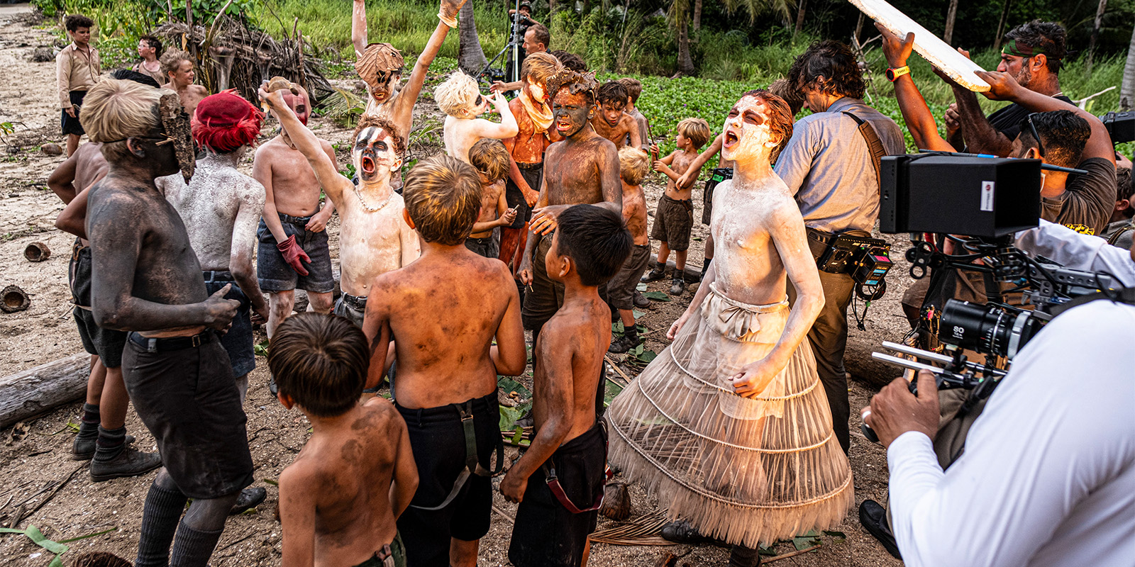 A group of children with painted faces and bodies, some in tattered clothes, gather outdoors on a dirt ground. A film crew with camera equipment captures the scene; trees and greenery are in the background.