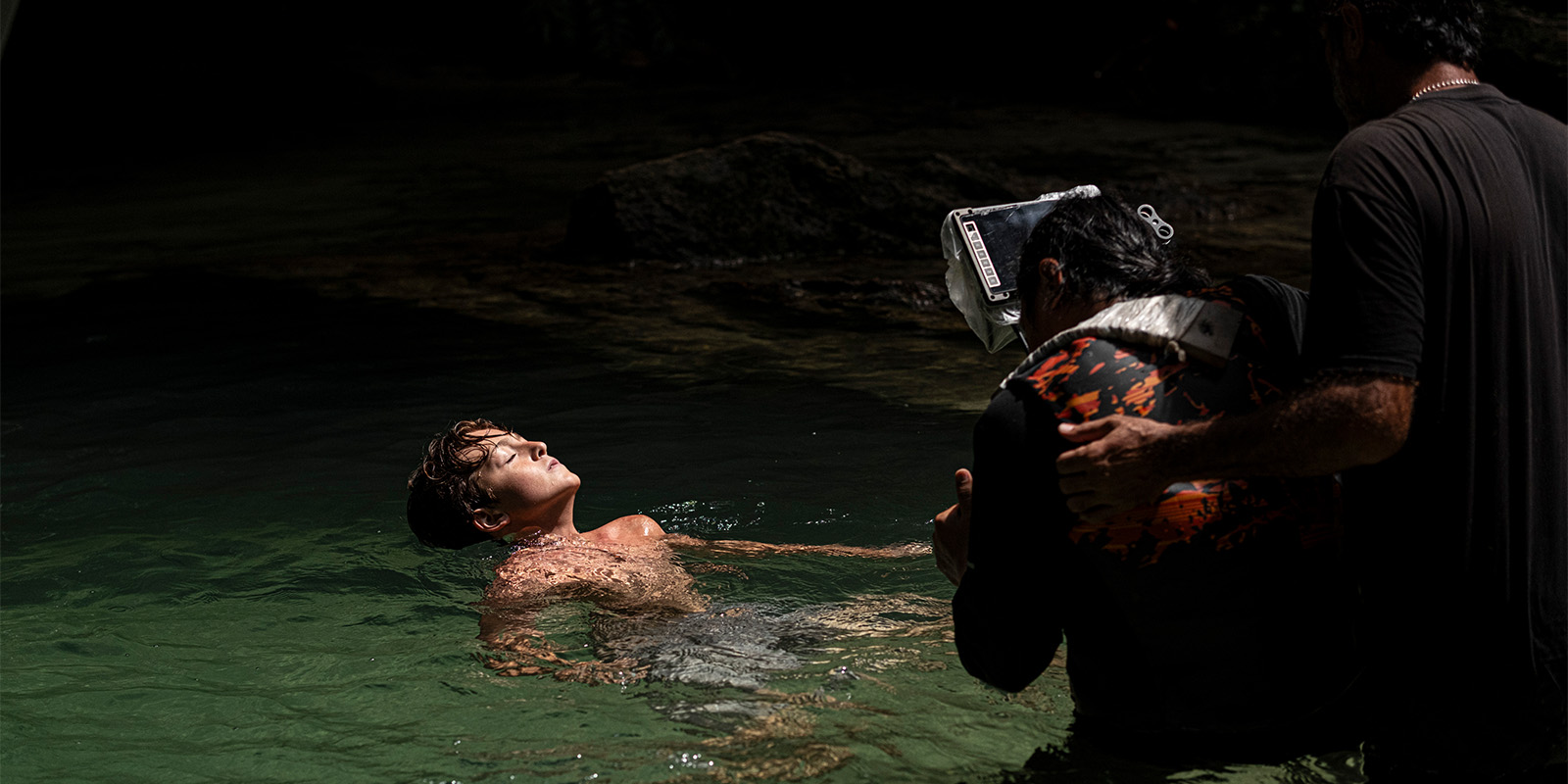 A young person floats in clear water with eyes closed, while two adults wearing dark shirts and one in a snorkel mask assist nearby. Sunlight highlights the youth, and the background is dim.