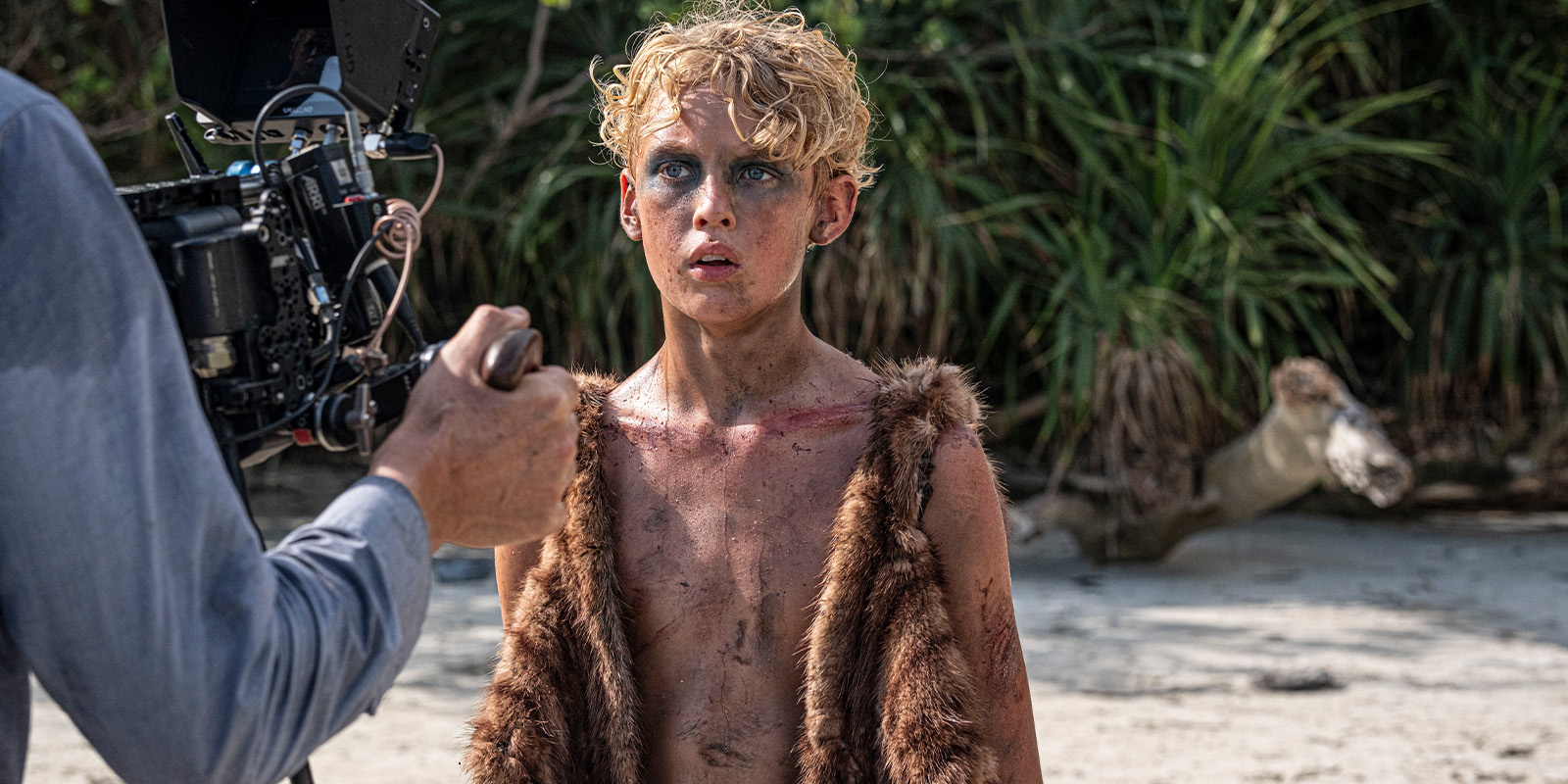 A camera operator films a young person with messy blond hair and face paint, wearing a fur waistcoat and standing bare-chested on a sandy beach, with greenery in the background.
