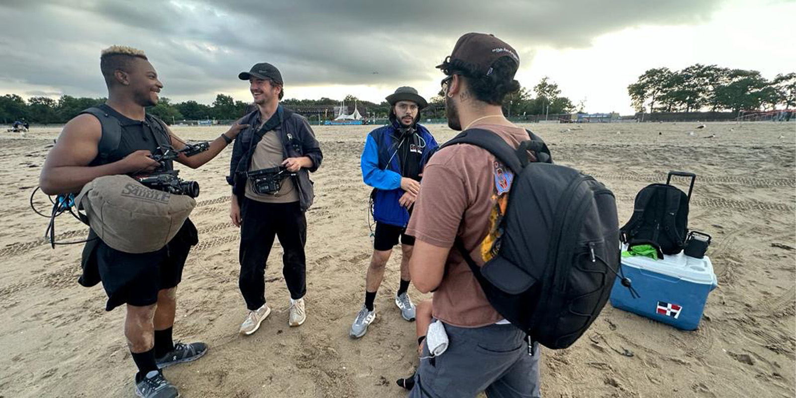 Four men stand on a sandy beach having a conversation; two hold camera equipment, one wears a blue jacket, and one has a rucksack. A cool box and more bags sit on the sand, with cloudy skies and trees in the background.