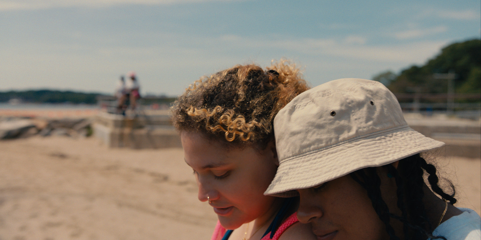 Two people sit close together on a sandy beach. One has curly hair and the other wears a light-coloured bucket hat. Both appear relaxed, with blurred figures and trees in the background.