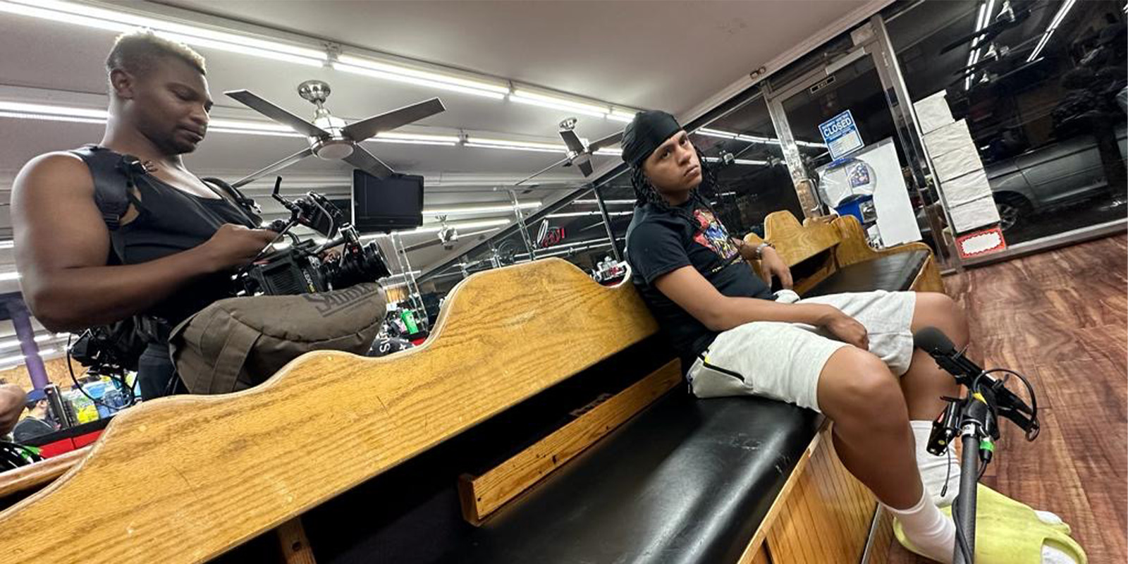 Two people are inside a barbershop. One person sits on a bench, looking at the camera, while the other stands beside him holding a camera and equipment. The shop has wooden benches and mirrors along the walls.