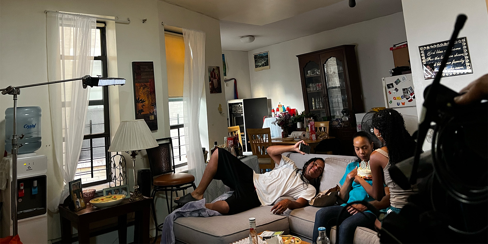 A man reclines on a sofa using his mobile while two women sit beside him, talking and eating. The room has a dining area, water cooler, fridge, and various household items. Film equipment is visible on the left.