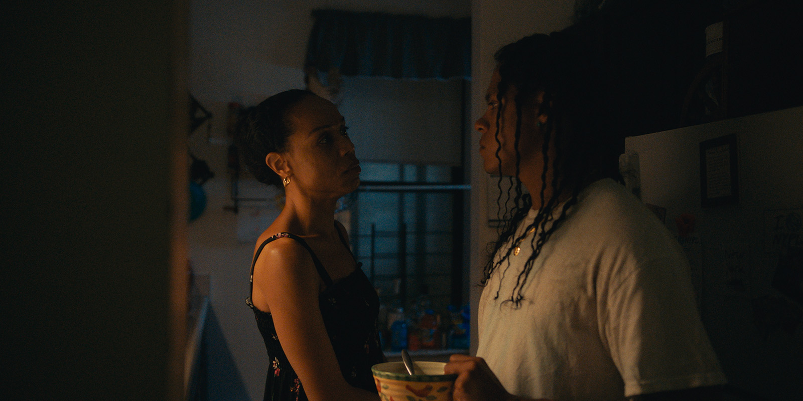 Two people stand close together in a dimly lit kitchen. The woman faces the man and looks at him seriously, while the man, holding a bowl and spoon, looks back at her. Light from a window partially illuminates the scene.