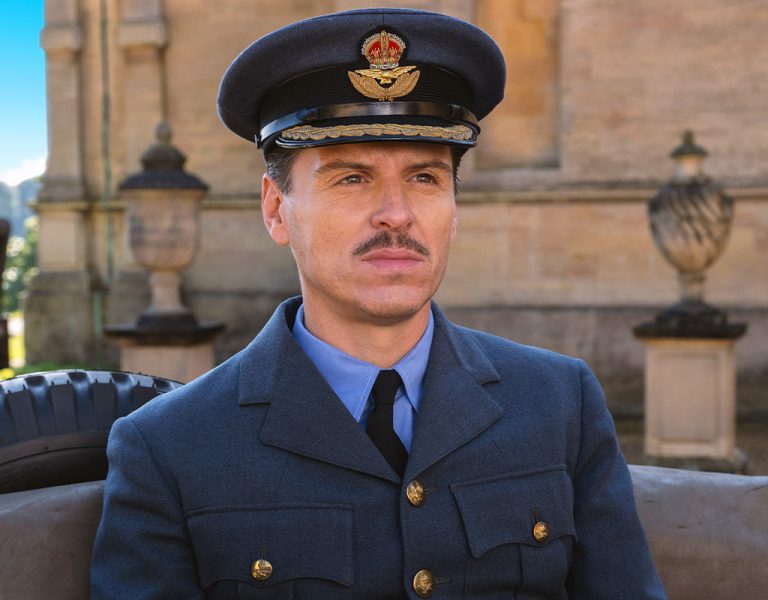 A man in a blue military uniform and cap with an aviation emblem sits outdoors in front of a stone building, with part of a tyre and a vintage vehicle visible behind him.