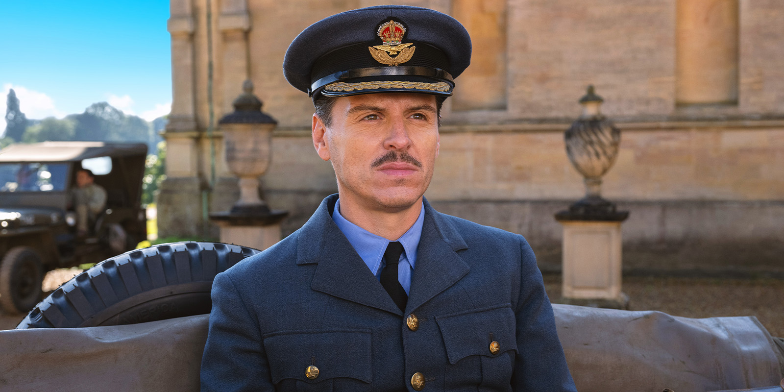 A man in a blue military uniform and cap with an aviation emblem sits outdoors in front of a stone building, with part of a tyre and a vintage vehicle visible behind him.