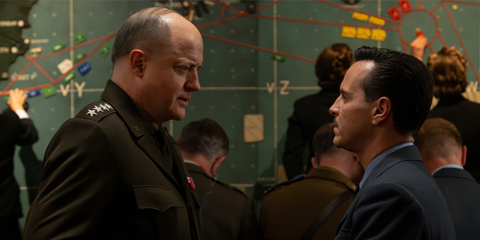 Two men, one in military uniform and one in a suit, stand facing each other and talking in a command centre with a large map on the wall and several people working in the background.