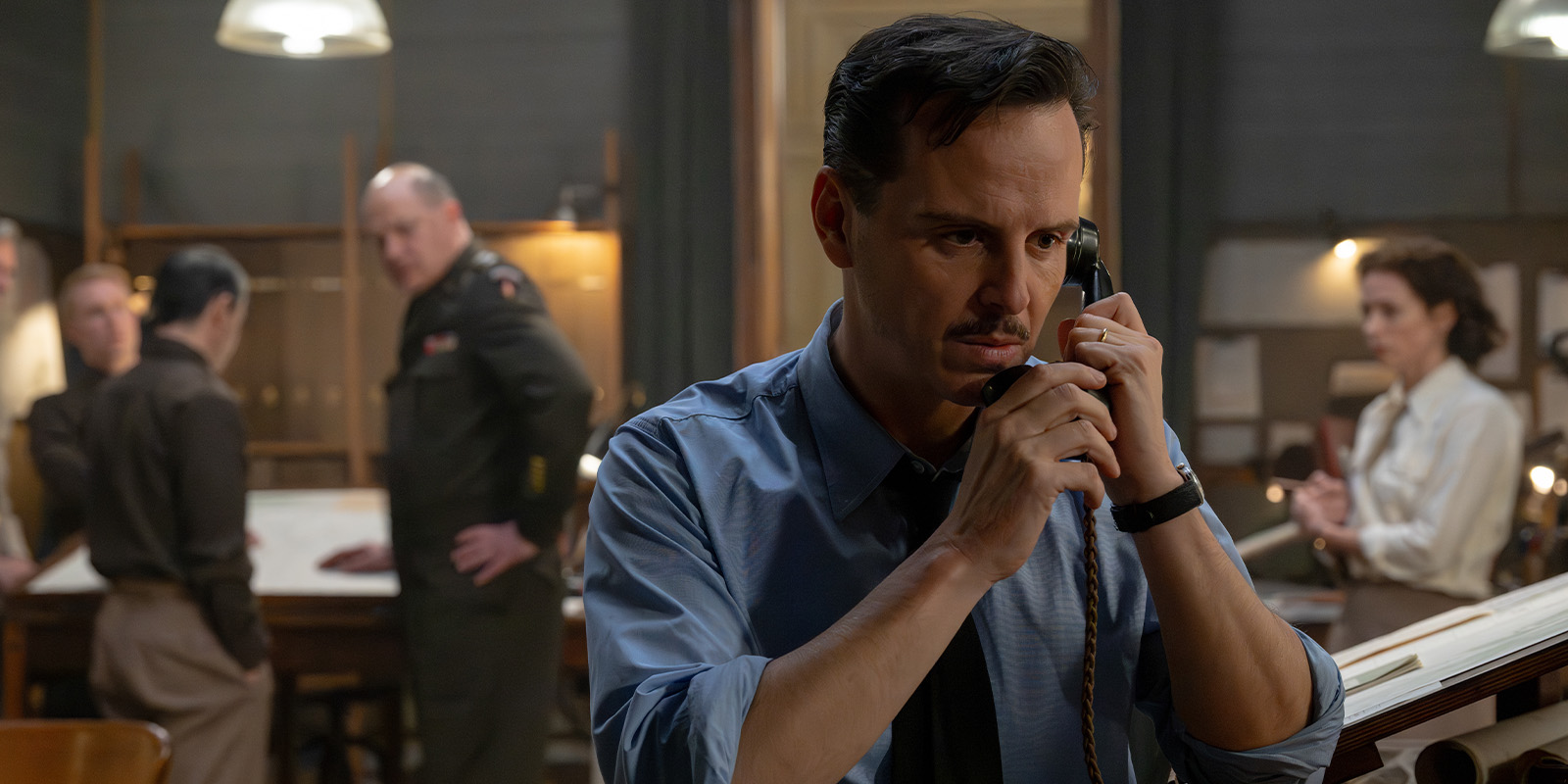 A man in a blue shirt holds a telephone receiver to his ear in a busy office. In the background, four people in military and civilian attire are gathered around a table, appearing to discuss documents.