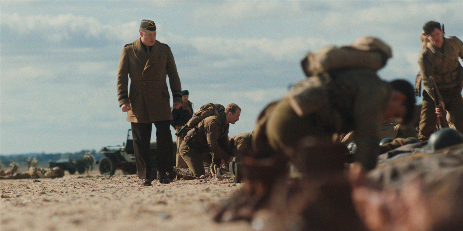 A group of soldiers in uniform are crouched on a sandy beach, some lying down, whilst one stands and walks nearby; vehicles are visible in the background under a partly cloudy sky.