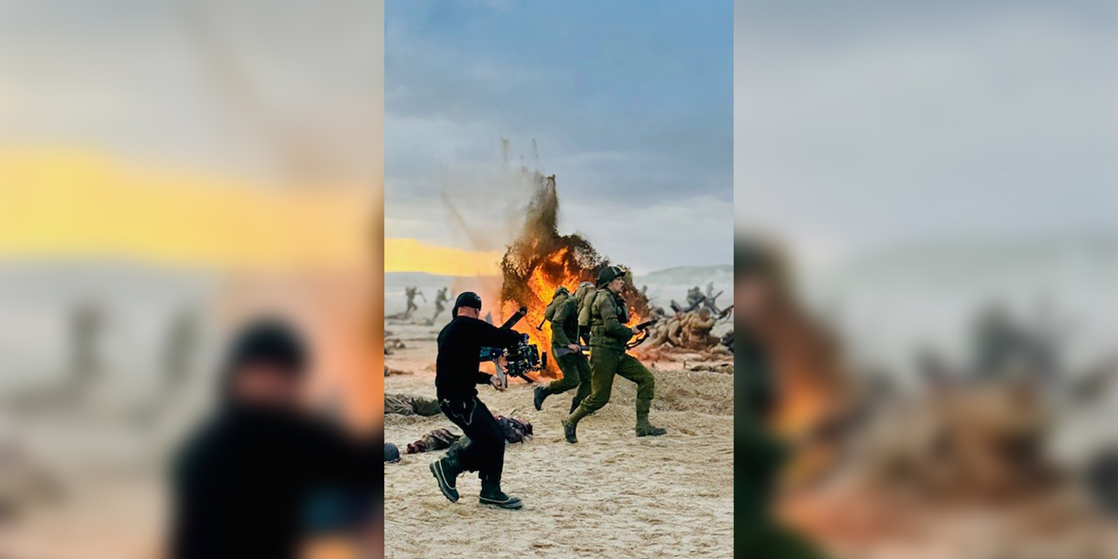 A cameraman films a person in military gear running past a large fire outdoors, with scattered debris and other figures visible in the background.
