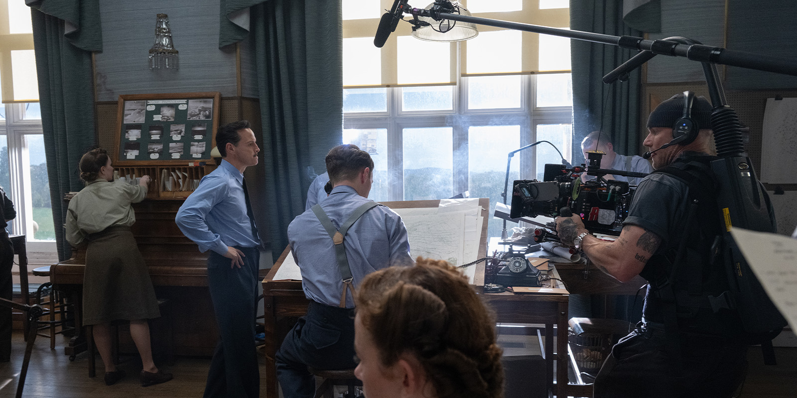 A film crew films a scene in a room with large windows. Actors in period clothing stand and sit at desks with papers and equipment while a camera operator and crew film them. A boom microphone is positioned above.