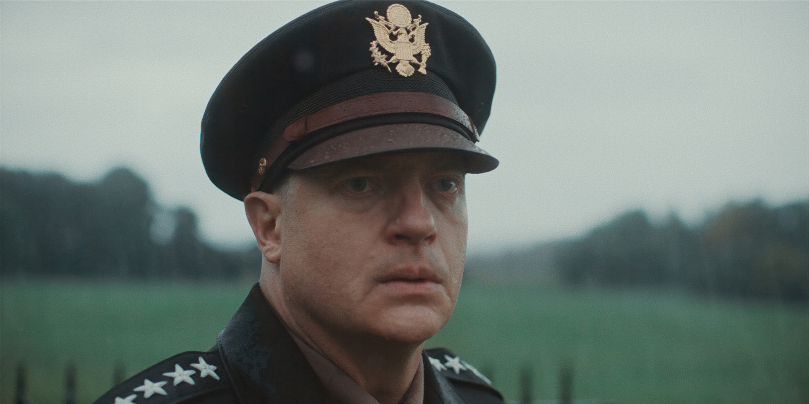 A man wearing a decorated military uniform and cap stands outdoors on a rainy day, with a blurred green field and trees in the background.