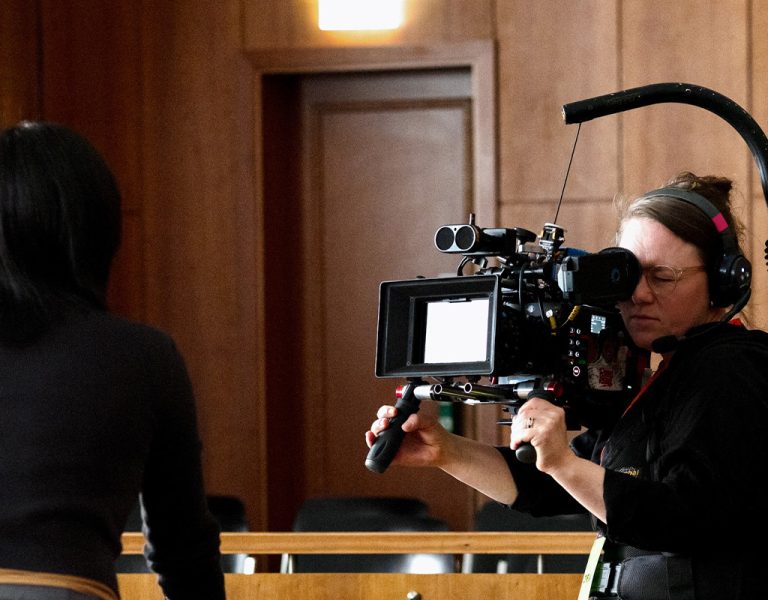 A person operating a professional video camera films another person in an indoor setting with wood-panelled walls and empty seats in the background.