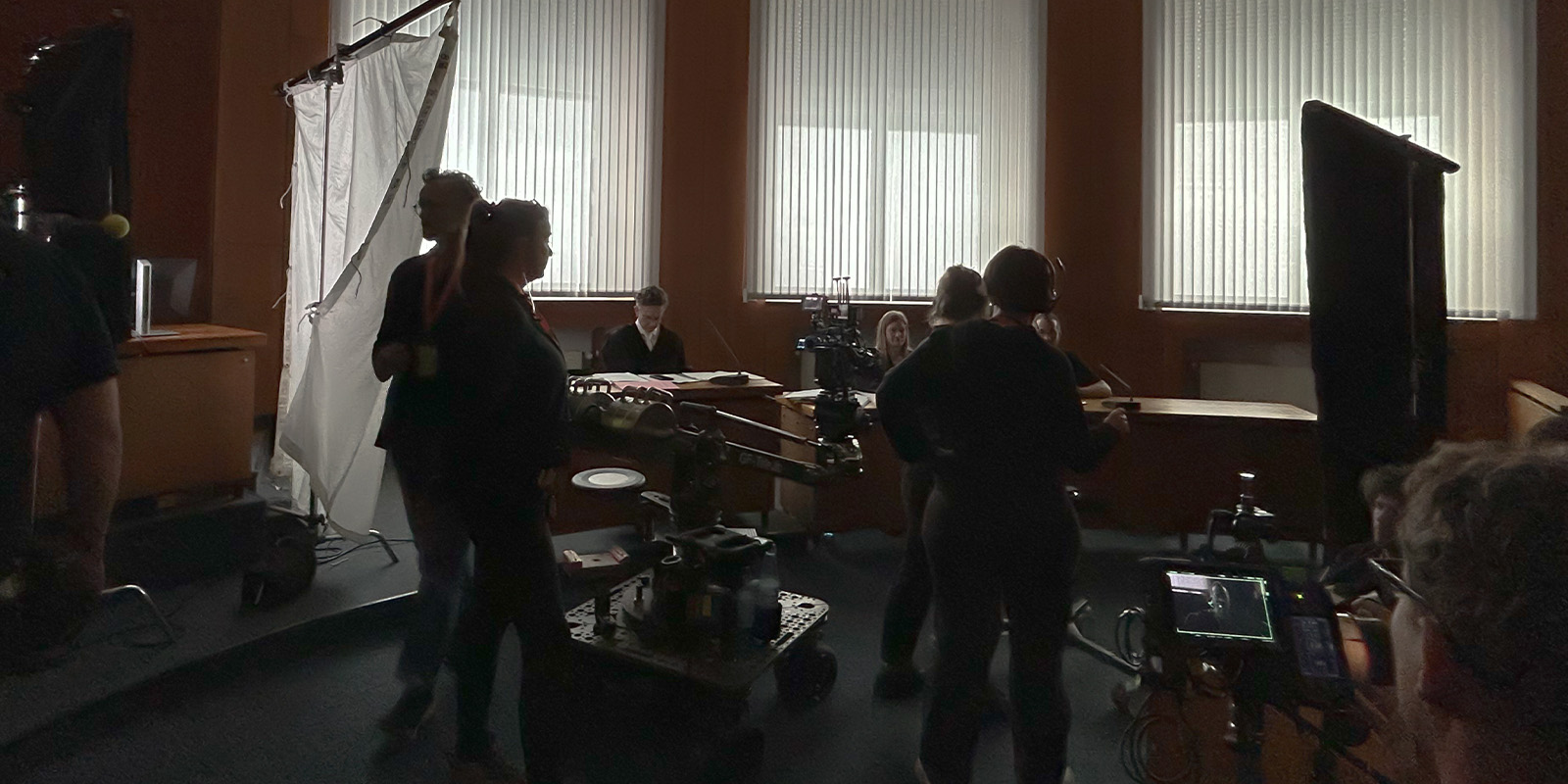 People are working on a film set indoors with cameras, lighting equipment, and monitors visible. Several individuals are silhouetted against large windows with vertical blinds. The environment appears busy and professional.