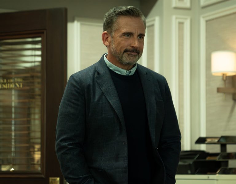 A man with short hair and a beard, wearing a dark blazer over a jumper and collared shirt, stands in an office with white panelled walls, a lamp, and a glass door labelled President.