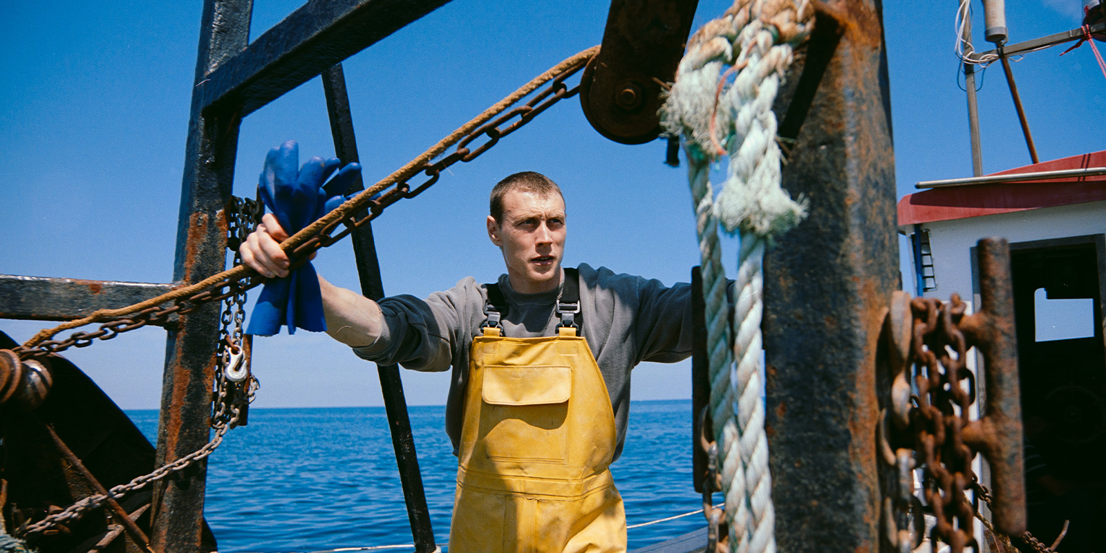A person wearing yellow waterproof overalls and holding blue gloves stands on a fishing boat surrounded by chains, ropes, and metal structures, with the sea and clear sky in the background.