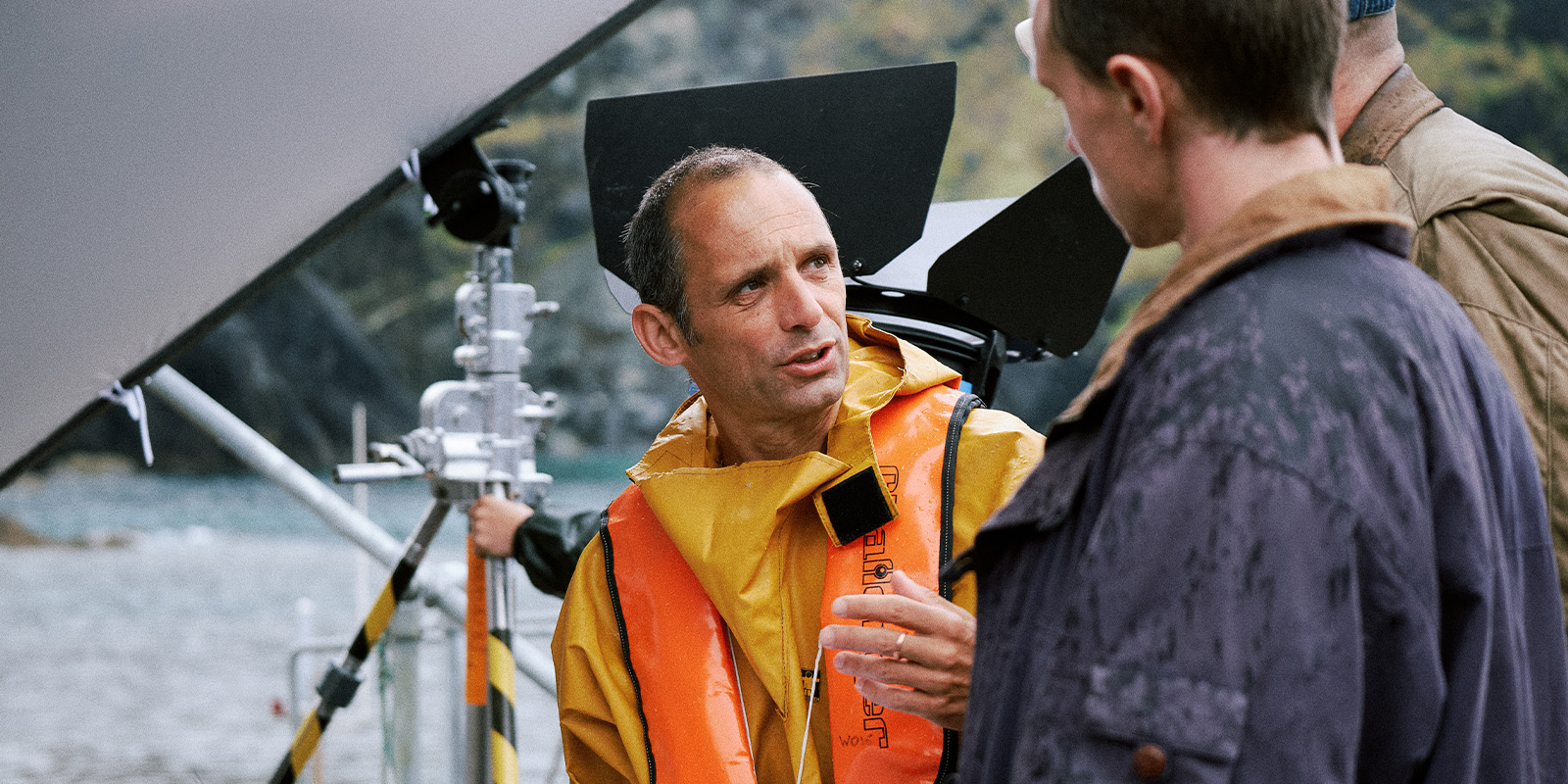 A person wearing a yellow raincoat and orange safety vest talks to another person outdoors near film production equipment, with water and hills visible in the background.