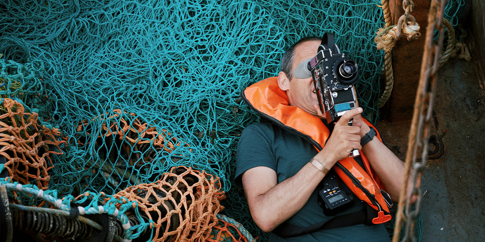 A person wearing an orange life jacket and holding a vintage film camera sits among a pile of fishing nets.