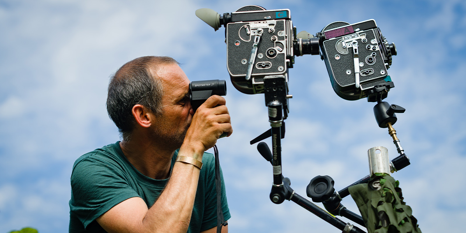 A person wearing a green shirt looks through a small handheld device near two vintage film cameras mounted on adjustable stands outdoors against a partly cloudy sky.