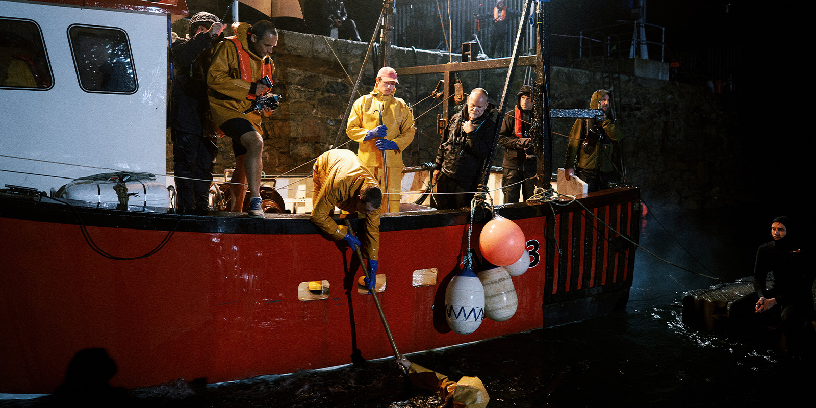 Several people in yellow rain gear and others in dark clothing stand on a red and white fishing boat at night. One person leans over the side with a net, near floating buoys and barrels. The scene is lit by artificial light.
