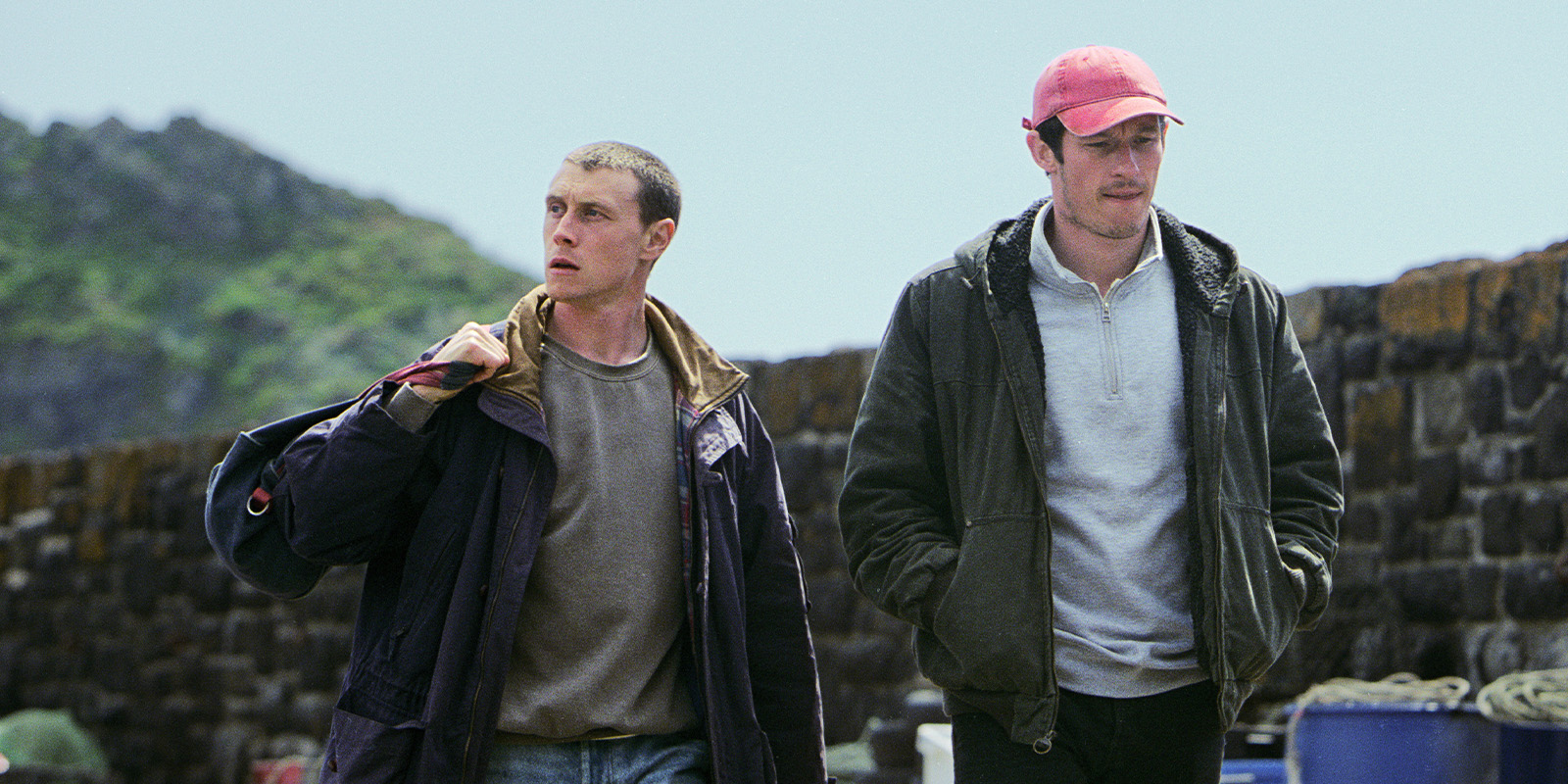 Two men walk outdoors by a stone wall; one carries a duffle bag over his shoulder, the other wears a red cap and has his hands in his pockets. There is greenery and a hill in the background.