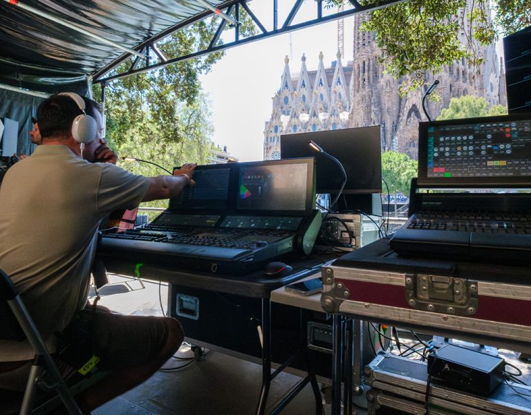 A person wearing headphones sits at a control panel with multiple screens and audio equipment under a canopy. The Sagrada Família basilica is visible in the background outside.