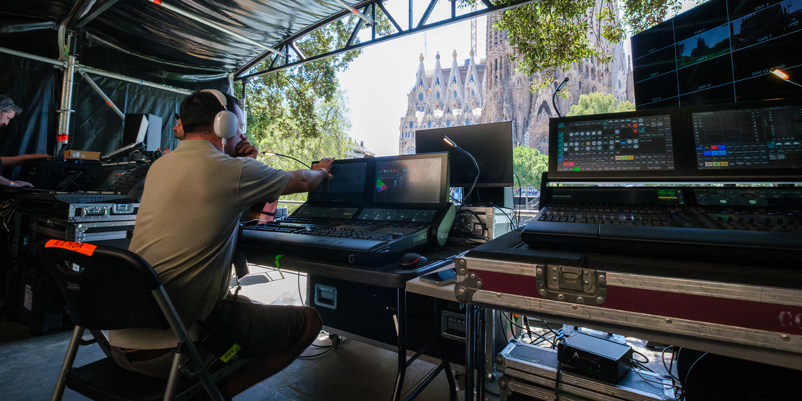 A person wearing headphones sits at a control panel with multiple screens and audio equipment under a canopy. The Sagrada Família basilica is visible in the background outside.