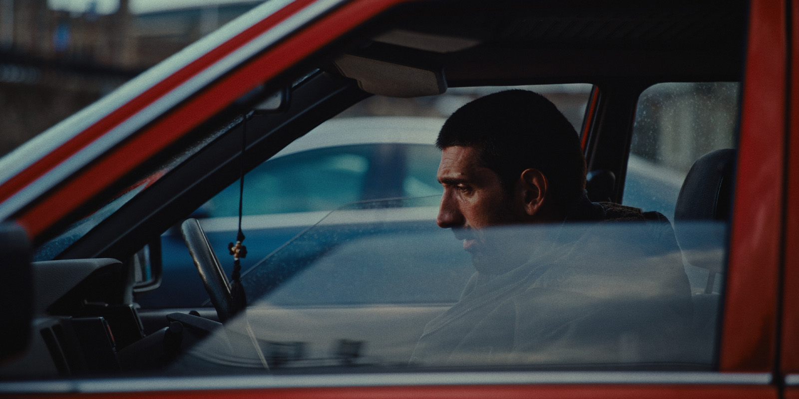 A man sits in the driver’s seat of a red car, looking forward with a serious expression. The photo is taken from outside the car, with reflections on the window partially obscuring his face.