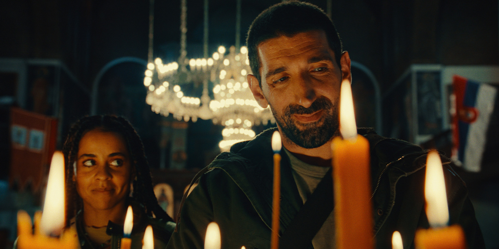 Two people stand close together in front of lit candles. The man looks down at the candles, whilst the woman, standing beside him, looks at him and smiles. A chandelier with more lights hangs in the background.