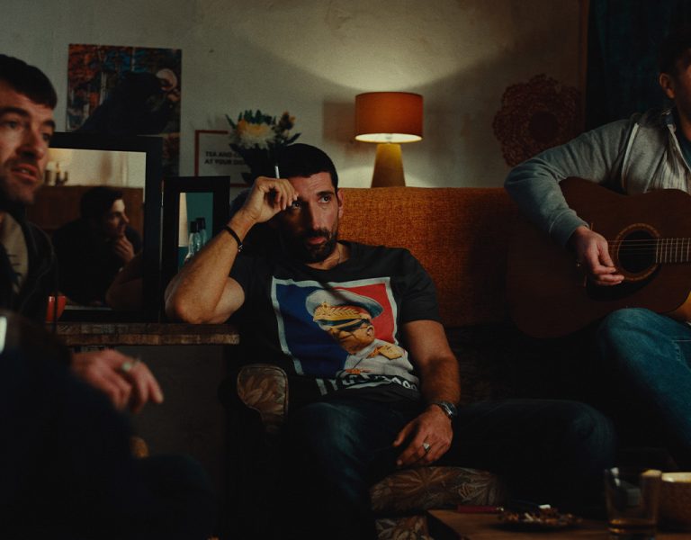 Three men sit in a dimly lit sitting room. One plays an acoustic guitar, another rests his hand on his face, and the third looks ahead. Various lamps, artwork, and snacks are visible in the background.