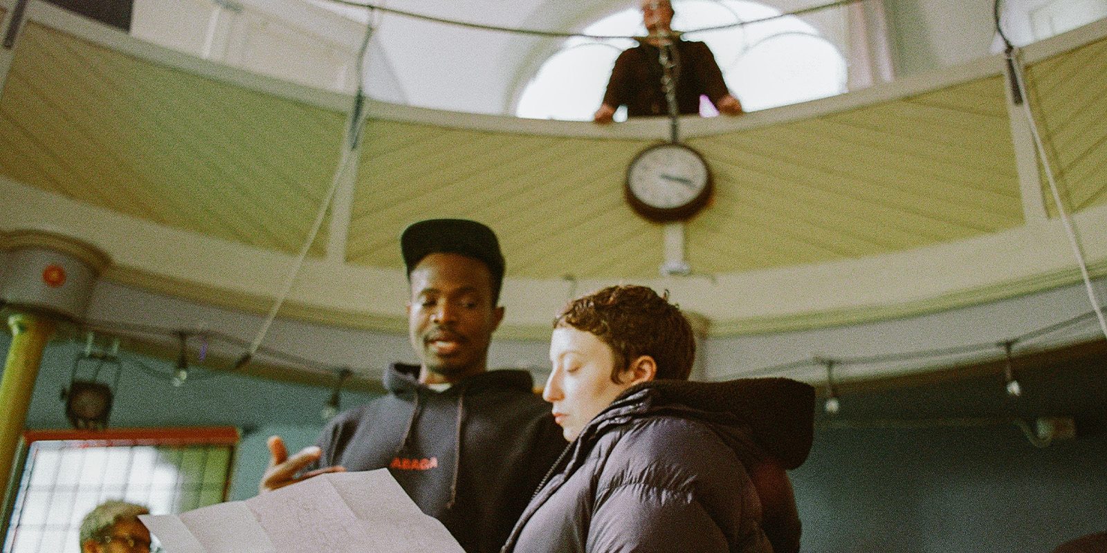 Two people stand together indoors, looking at a sheet of paper, while a third person stands on an upper level balcony above them, near a clock mounted on the wall. The setting appears to be a public or community space.