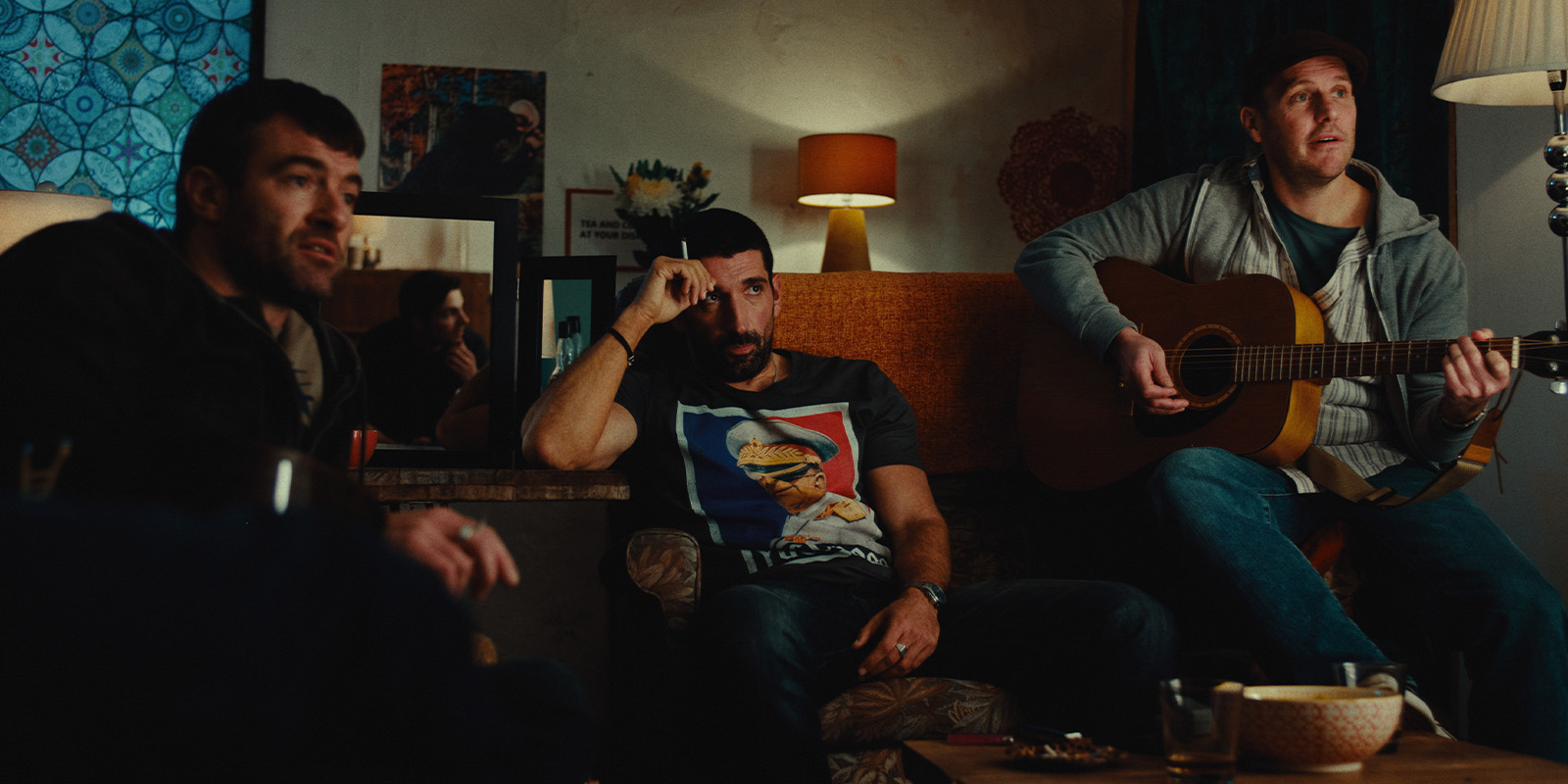 Three men sit in a dimly lit sitting room. One plays an acoustic guitar, another rests his hand on his face, and the third looks ahead. Various lamps, artwork, and snacks are visible in the background.