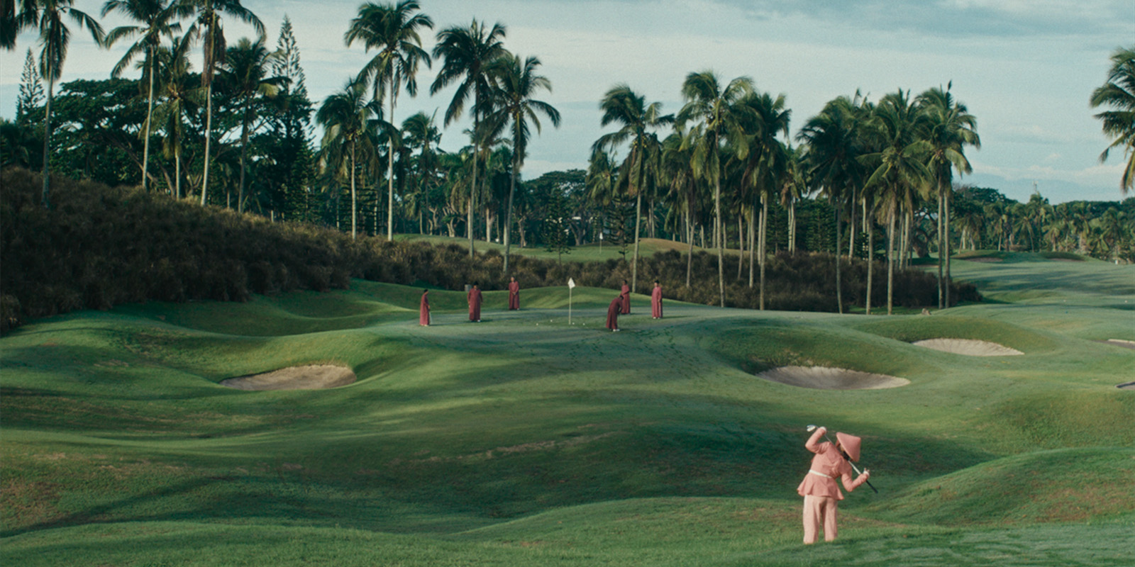 A group of people in red robes stand on a green golf course surrounded by palm trees, whilst a person in a pink elephant costume stands in the foreground facing away.