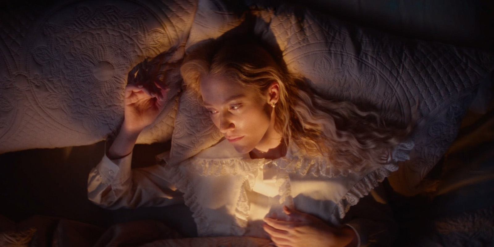 A person with long, wavy blonde hair lies on a bed, looking up thoughtfully. They wear a light-coloured, ruffled shirt and are illuminated by warm, soft lighting. The bedspread and pillows have a quilted pattern.