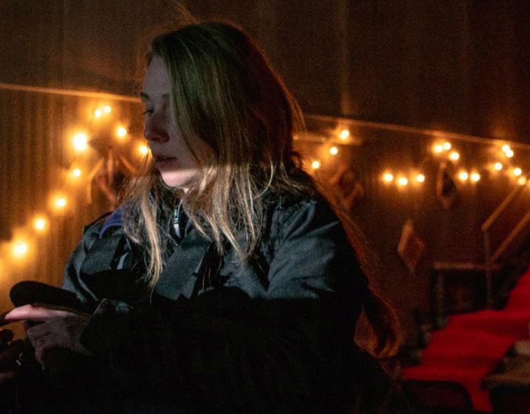 A woman in a dark room holds a torch and looks at a device. Warm fairy lights and a red-carpeted staircase are visible in the background.