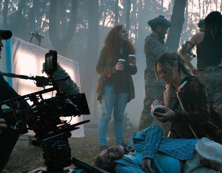 A film crew works on an outdoor set in a forest. A camera operator films an actor lying on the ground while another actor kneels by, holding a prop heart. Crew members stand in the background holding drinks.