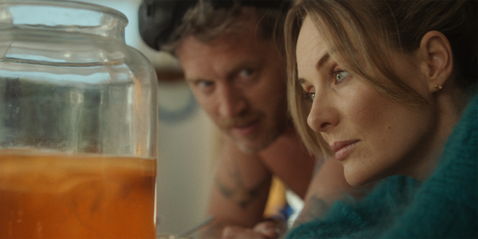 A woman with light brown hair in a teal jumper looks thoughtfully at a large glass jar of orange liquid, while a man in goggles and a vest looks on from behind her.