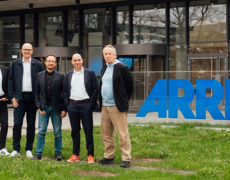 Five men stand in front of a modern glass building with large blue ARRI logos. The men are dressed in business casual attire and stand on a paved walkway bordered by grass.