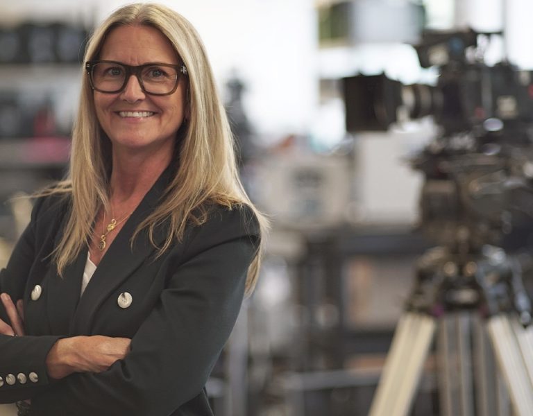 Dana Harrison with arms folded, smiling into camera with a large camera behind