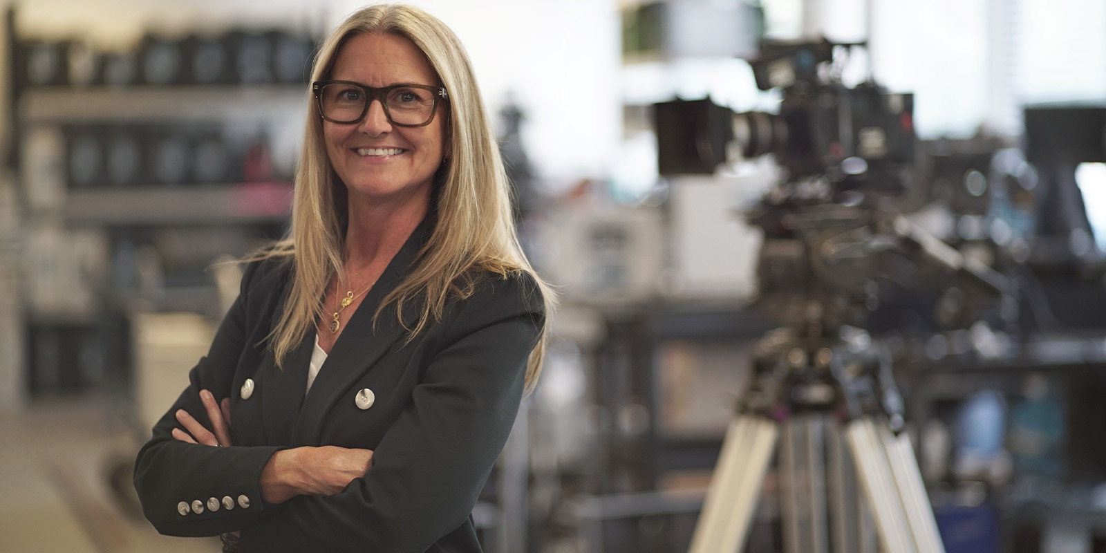 Dana Harrison with arms folded, smiling into camera with a large camera behind