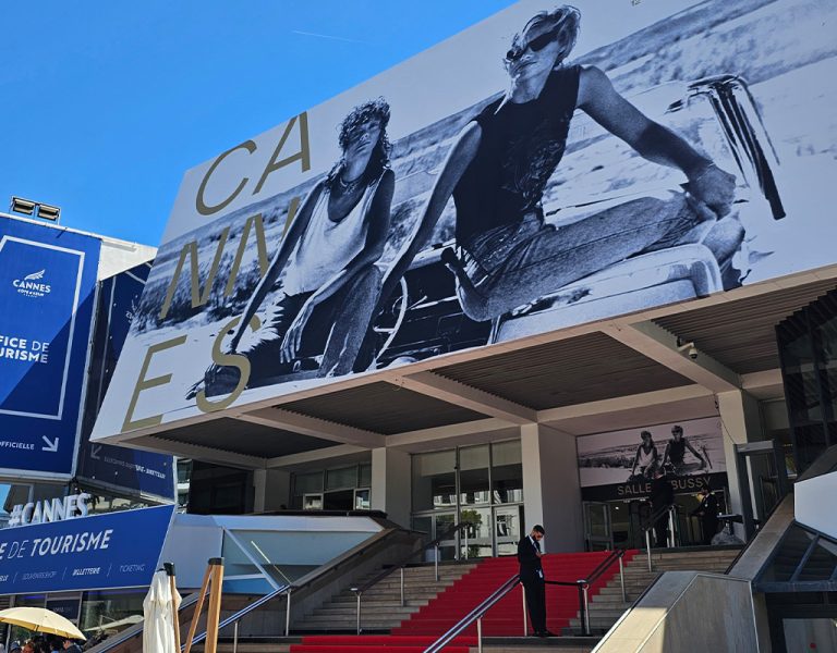 Large black-and-white hoarding of two people riding bicycles above the Cannes Film Festival entrance, featuring red carpeted stairs, blue signage for the tourist office, and people walking below on a sunny day.