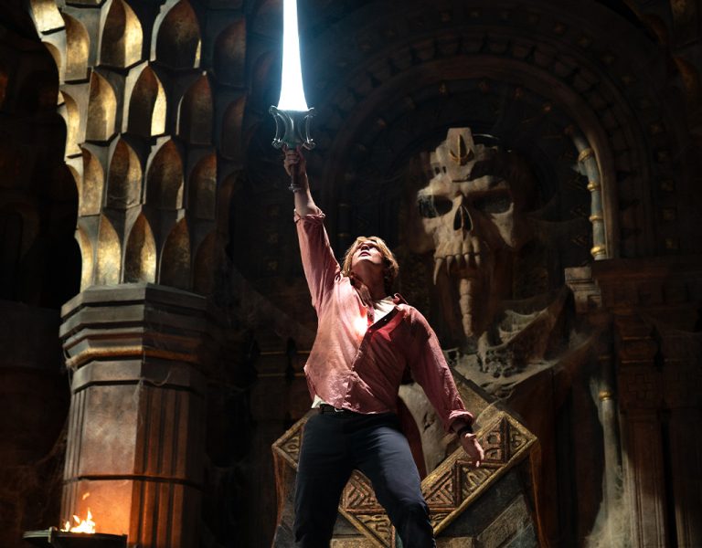 A person stands on a raised platform in a dim, ornate chamber, holding a glowing sword aloft. A large skeletal face is carved into the stone wall behind them. Fire torches flank the scene.