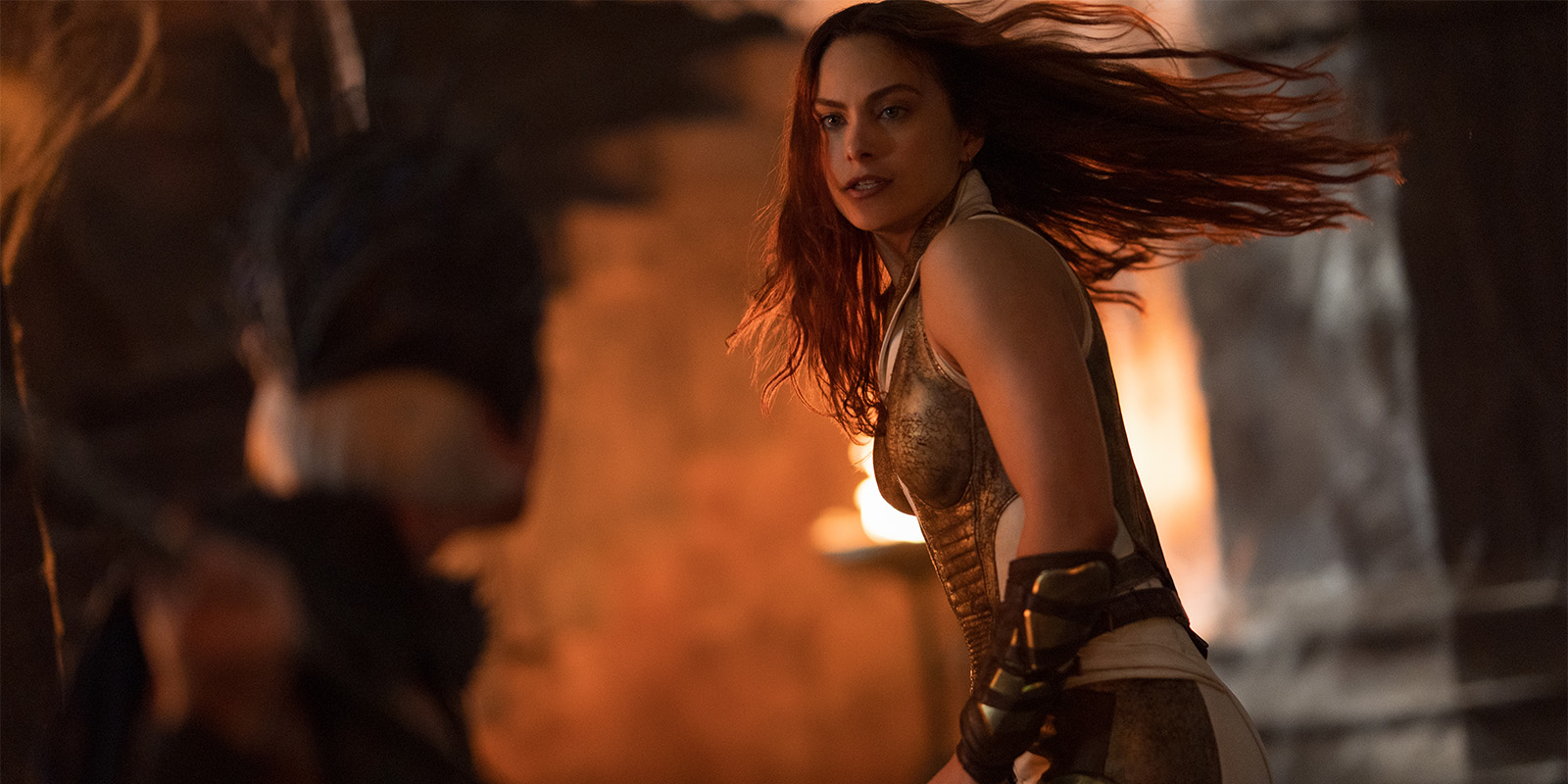 A woman in gold armour with long red hair stands in a dramatic, dimly lit scene, turning towards another figure. The background is blurred, with hints of fire and stone walls visible.