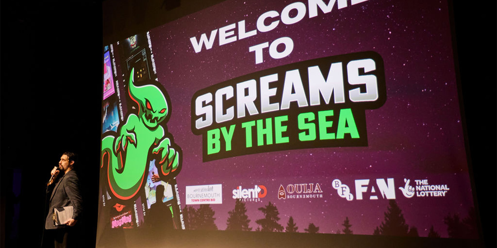A person presenting in front of the Screams By The Sea poster