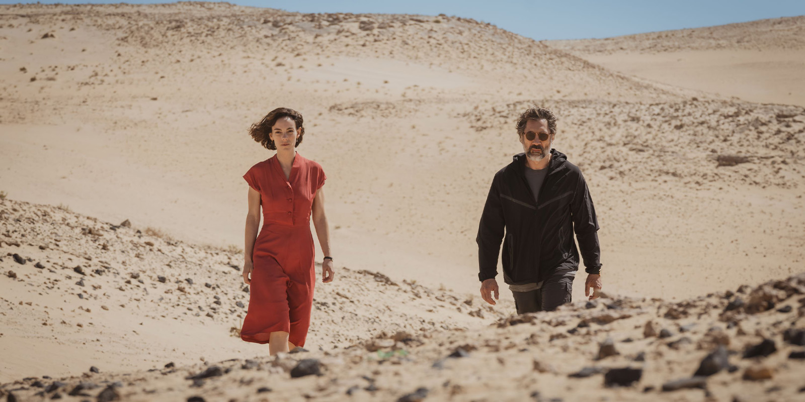 A woman in a red dress and a man in dark clothing walk through a rocky desert landscape under a clear blue sky.