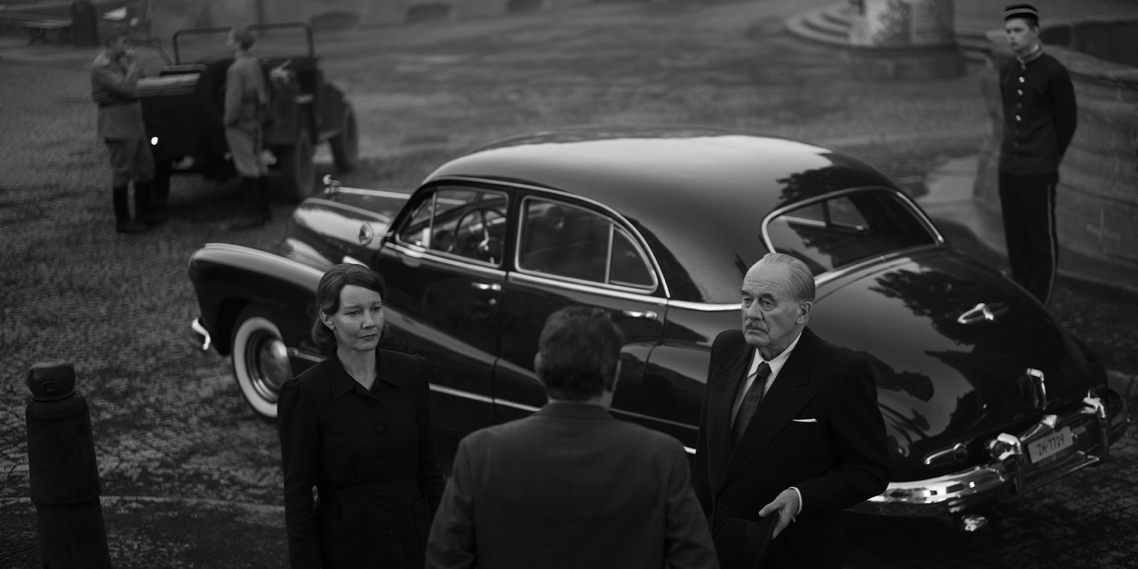 Two men in suits and a woman stand near a vintage car on a cobbled street. Two uniformed guards and an open-top vehicle are in the background. The image is in black and white.